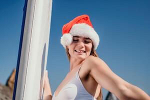 femme mer souper. proche en haut portrait de content Jeune caucasien femme avec longue cheveux dans Père Noël chapeau à la recherche à caméra et souriant. mignonne femme portrait dans une blanc bikini posant sur souper planche dans le mer photo