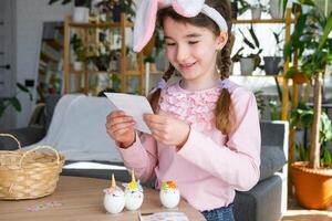 une mignonne fille avec rose lapin oreilles fait du un Pâques artisanat - décore un Oeuf dans le forme de une Licorne avec strass, corne, fleurs dans le intérieur de une maison avec les plantes. photo