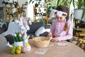 une mignonne fille avec rose lapin oreilles fait du un Pâques artisanat - décore un Oeuf dans le forme de une Licorne avec strass, corne, fleurs dans le intérieur de une maison avec les plantes. photo