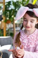 une mignonne fille avec rose lapin oreilles fait du un Pâques artisanat - décore un Oeuf dans le forme de une Licorne avec strass, corne, fleurs dans le intérieur de une maison avec les plantes. photo