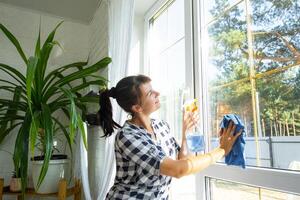 femme manuellement lavages le fenêtre de le maison avec une chiffon avec vaporisateur nettoyeur et balai à l'intérieur le intérieur avec blanc rideaux. restauration commande et propreté dans le printemps, nettoyage servir photo