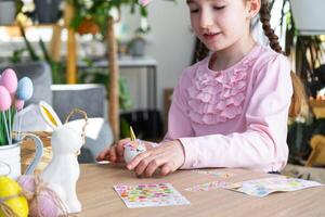 une mignonne fille avec rose lapin oreilles fait du un Pâques artisanat - décore un Oeuf dans le forme de une Licorne avec strass, corne, fleurs dans le intérieur de une maison avec les plantes. photo