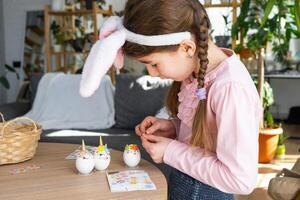 une mignonne fille avec rose lapin oreilles fait du un Pâques artisanat - décore un Oeuf dans le forme de une Licorne avec strass, corne, fleurs dans le intérieur de une maison avec les plantes. photo