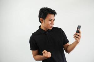 excité content Jeune asiatique homme gagnant sentiment joie en utilisant téléphone intelligent gagnant loterie jeu, pari, avoir en espèces retour en ligne cadeau dans mobile app message en portant cellule téléphone isolé sur blanc Contexte. photo