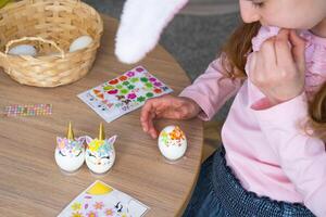 une mignonne fille avec rose lapin oreilles fait du un Pâques artisanat - décore un Oeuf dans le forme de une Licorne avec strass, corne, fleurs dans le intérieur de une maison avec les plantes. photo