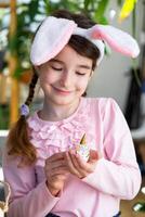 une mignonne fille avec rose lapin oreilles fait du un Pâques artisanat - décore un Oeuf dans le forme de une Licorne avec strass, corne, fleurs dans le intérieur de une maison avec les plantes. photo