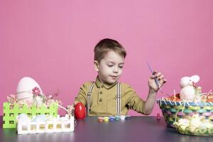 charmant enfant avec une sourire artisanat des œufs et décorations pour le Pâques festivités, en utilisant art Provisions et aquarelle peindre. peu heureux garçon sentiment excité à propos La peinture activité. photo