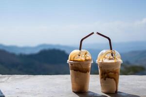 deux tasses de glacé café avec crémeux garnitures et caramel sirop à la perfection complété par le expansif vue de une Montagne paysage horizon jouissance Voyage vacances photo