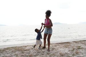 mixte course africain et asiatique mère et garçon est en jouant à le Extérieur zone. souriant content famille avoir amusement fonctionnement sur le plage. portrait de maman et enfant mode de vie avec une unique coiffure. photo