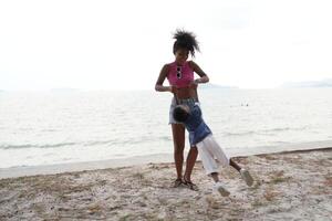 mixte course africain et asiatique mère et garçon est en jouant à le Extérieur zone. souriant content famille avoir amusement fonctionnement sur le plage. portrait de maman et enfant mode de vie avec une unique coiffure. photo