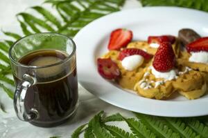 gaufres avec fraise, une tasse de café et vert feuilles de fougère sur une tableau. magnifique et savoureux petit déjeuner. photo