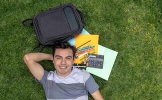 une Masculin étudiant mensonge sur le herbe avec une sac à dos et école Provisions photo