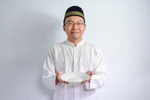 asiatique musulman homme portant des lunettes et blanc tissu tenir vide assiette pour jeûne ramadan. isolé blanc Contexte photo