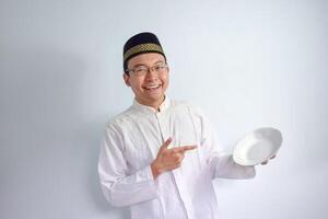 asiatique musulman homme portant des lunettes et blanc tissu montrer du doigt vide assiette pour jeûne ramadan. isolé blanc Contexte photo