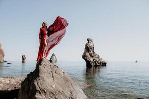 femme Voyage mer. Jeune content femme dans une longue rouge robe posant sur une plage près le mer sur Contexte de volcanique rochers, comme dans Islande, partage Voyage aventure périple photo