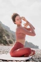 milieu vieilli bien à la recherche femme avec noir cheveux, aptitude instructeur dans leggings et hauts Faire élongation et pilates sur yoga tapis près le mer. femelle aptitude yoga routine concept. en bonne santé mode de vie photo