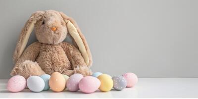 ai généré une photo avec une peluche marron lapin jouet avec longue oreilles séance dans de face de une rangée de coloré peint des œufs. le lapin apparaît mignonne et duveteux contre le vibrant toile de fond de le des œufs