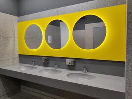 moderne salle de bains intérieur avec pierre gris carrelage, évier et et rond Jaune miroirs. salle de bains avec lavabo et robinet. Publique salle de bains dans le aéroport ou restaurant, café, Bureau photo
