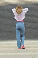 une blond fille dans jeans et une blanc chandail des stands avec sa retour à le caméra contre une gris brique mur. photo
