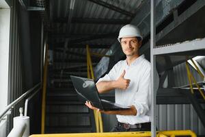 Jeune souriant professionnel dans salopette et protecteur casque permanent dans de face de caméra à l'intérieur grand machine de construction plante photo