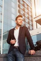 Beau Jeune homme d'affaire avec une barbe et dans une affaires costume permanent sur le rue contre le Contexte de le Bureau bâtiment suivant à une confortable élégant cuir sac. photo