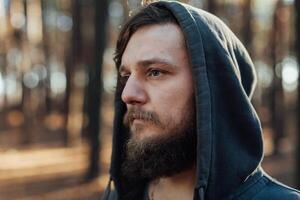 une Jeune homme avec une barbe des promenades dans une pin forêt. portrait de une brutal barbu homme dans une capuche photo