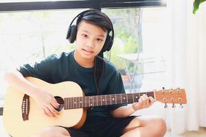 asiatique garçon portant écouteurs avec noir la musique asseoir et jouer guitare dans le maison. la musique apprentissage concept, la musique compétence entraînement. photo