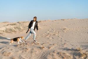 une Jeune caucasien fille quelque part dans une cuir veste et bleu jeans court le long de le sablonneux plage avec sa beagle chien photo