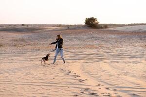 une Jeune caucasien fille quelque part dans une cuir veste et bleu jeans court le long de le sablonneux plage avec sa beagle chien photo