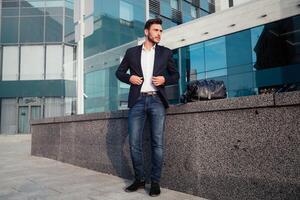 Beau Jeune homme d'affaire avec une barbe et dans une affaires costume permanent sur le rue contre le Contexte de le Bureau bâtiment suivant à une confortable élégant cuir sac. photo