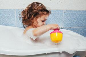 magnifique peu fille prise une une baignoire à maison. une mignonne bébé est séance dans le salle de bains et en jouant avec jouets et l'eau. personnel hygiène pour les enfants. du quotidien hygiène photo