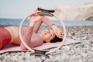 femme mer pilates. sportif content milieu vieilli femme pratiquant aptitude sur plage près mer, souriant actif femelle formation avec bague sur yoga tapis dehors, profiter en bonne santé mode de vie, harmonie et méditation photo