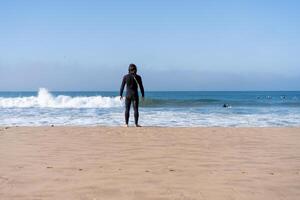 un homme habillé noir combinaison permanent océan plage arrière vue à la recherche sur horizon photo