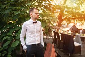 une Beau Jeune affaires homme dans une blanc chemise et attacher une papillon à une affaires dîner attend pour le sien collègues seul à une été restaurant table photo