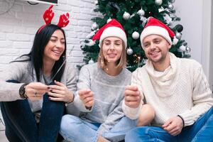 Jeune content couple dans Noël Chapeaux près une Noël arbre embrasser, en portant des lunettes de du vin. Nouveau année fête photo