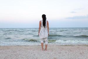 retour vue de magnifique femme dans blanc robe avec dreadlock sur le tête profiter le idyllique scène sur le plage. photo