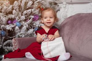 une magnifique mignonne peu fille habillé dans un élégant soir rouge robe est assis sur le canapé et ouvre une Nouveau années cadeau. avec Noël arbre sur le Contexte photo