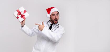 Jeune Beau médecin dans blanc uniforme et Père Noël claus chapeau permanent dans studio sur blanc Contexte sourire et spectacles une doigt à le cadeau boîte. photo