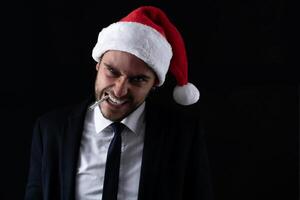 Jeune Beau caucasien gars dans affaires costume et Père Noël Chapeaux des stands sur noir Contexte dans studio avec sérieux visage et fumeur cigarette photo