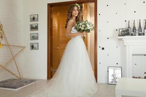 le la mariée dans une magnifique mariage robe avec une longue frisé cheveux des stands dans le chambre et regards à le caméra. la mariée à l'intérieur avec blanc fleurs bouquet et soumissionner couronne coiffure photo