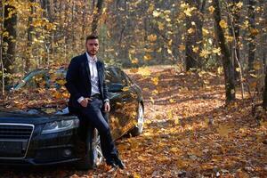 une Beau Jeune caucasien homme portant affaires costume permanent près noir voiture sur la nature. photo