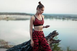 une magnifique brunette fille dans élégant des sports vêtements est repos après une sport actif faire des exercices près le lac. écoute à la musique sur écouteurs avec une téléphone intelligent dans mains photo