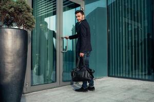 Beau Jeune homme d'affaire avec une barbe et dans une affaires costume permanent sur le rue contre le Contexte de le Bureau bâtiment suivant à une confortable élégant cuir sac. photo