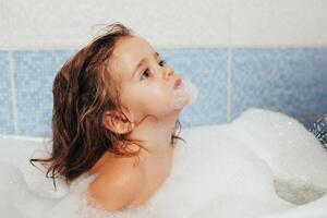 amusement de bonne humeur content bambin bébé prise une une baignoire en jouant avec mousse bulles. peu enfant dans une baignoire. souriant enfant dans salle de bains sur bleu Contexte. hygiène et santé se soucier. photo