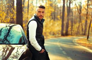 une Beau Jeune caucasien homme portant style noir gilet permanent près noir voiture sur la nature. photo