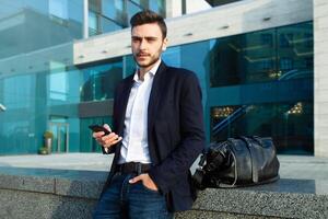 millénaire homme d'affaire avec une mobile téléphone dans le sien mains. Jeune réussi affaires élégant homme avec une noir cuir sac des promenades sur une ville rue contre le Contexte de Bureau bâtiments photo