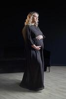 Enceinte femme posant dans un élégant noir robe à l'intérieur studio noir mur Contexte photo