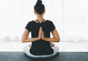 asiatique femmes Faire yoga méditer exercice à maison, séance dans lotus pose ou padmasana avec élevé mains namaste derrière le retour dans blanc chambre à coucher. travail en dehors portant noir chemise et blanc pantalon. photo