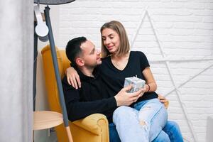 portrait de couple Jeune dans une confortable chambre. une homme est assis sur une Jaune chaise. une femme est assis sur le tour de une homme et détient une cadeau photo