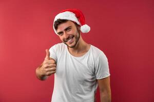 Jeune Beau caucasien gars dans une blanc T-shirt et Père Noël Chapeaux des stands sur rouge Contexte dans studio et montrant les pouces en haut. photo